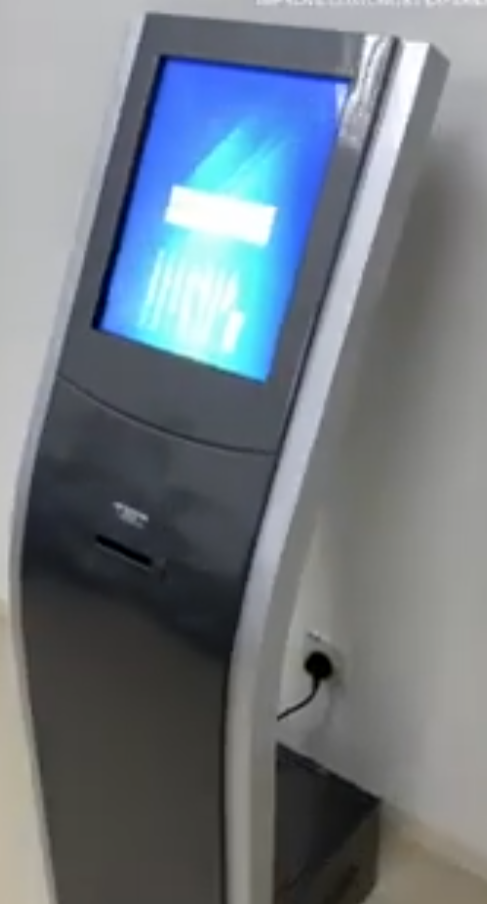 Free-standing queue ticket kiosk installed against a wall, with touchscreen and ticket outlet slot